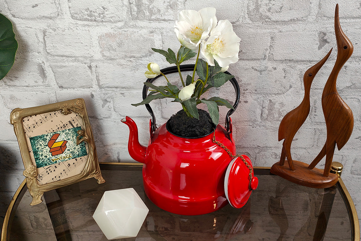 Arrangierte Vintage-Dekoration: roter Emaille-Wasserkessel mit weißen Blumen, Jugendstil-Bilderrahmen mit Musiknoten, Holzskulptur zweier Reiher.